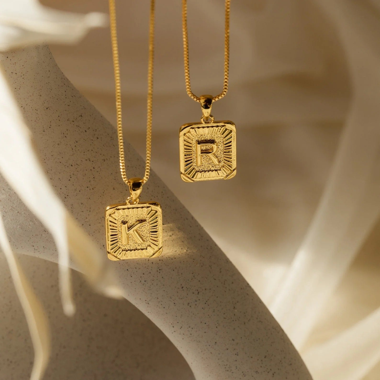 Two gold necklaces with square pendants displaying letters 'K' and 'R' on a neutral background.