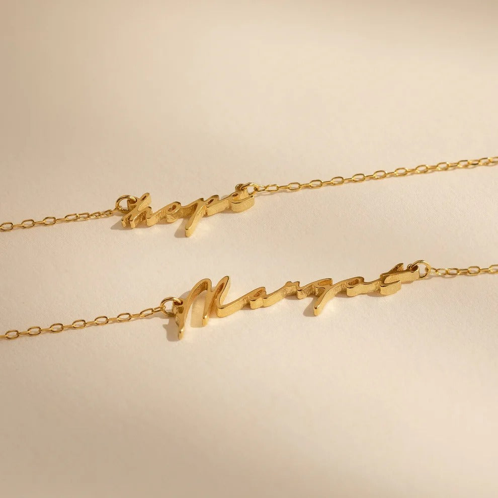 Two gold necklaces with script designs on a beige background