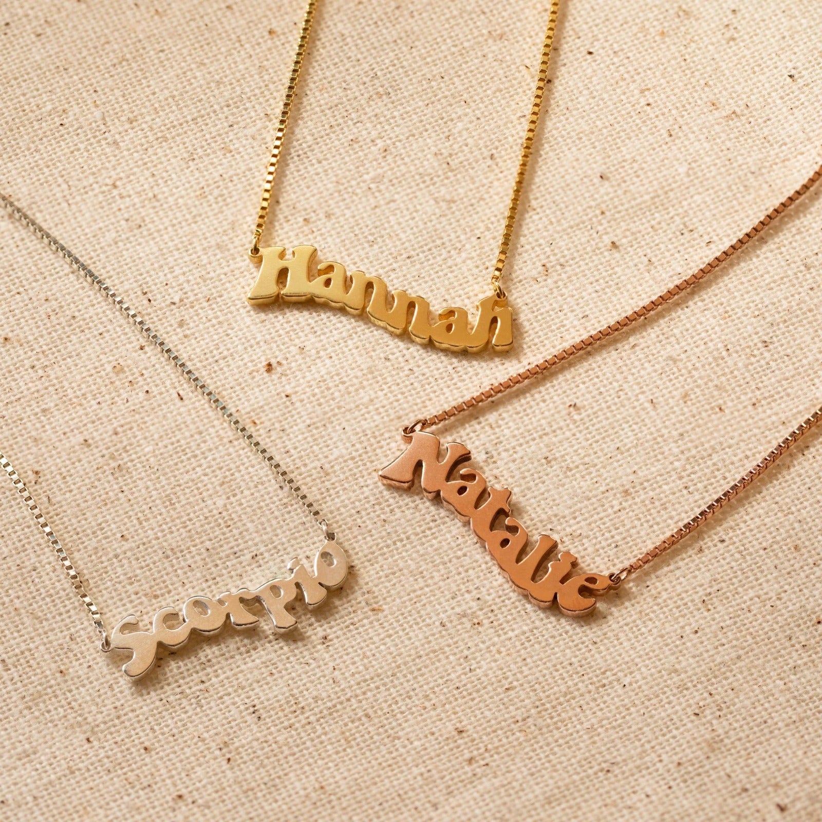 Three name necklaces in gold, silver, and rose gold on a beige fabric background.