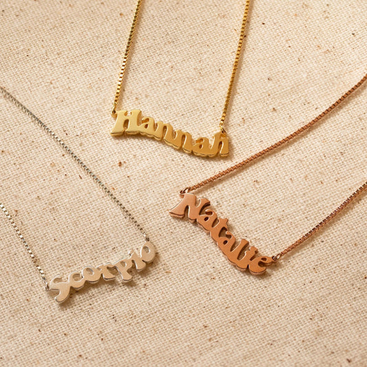 Three name necklaces in gold, silver, and rose gold on a beige fabric background.