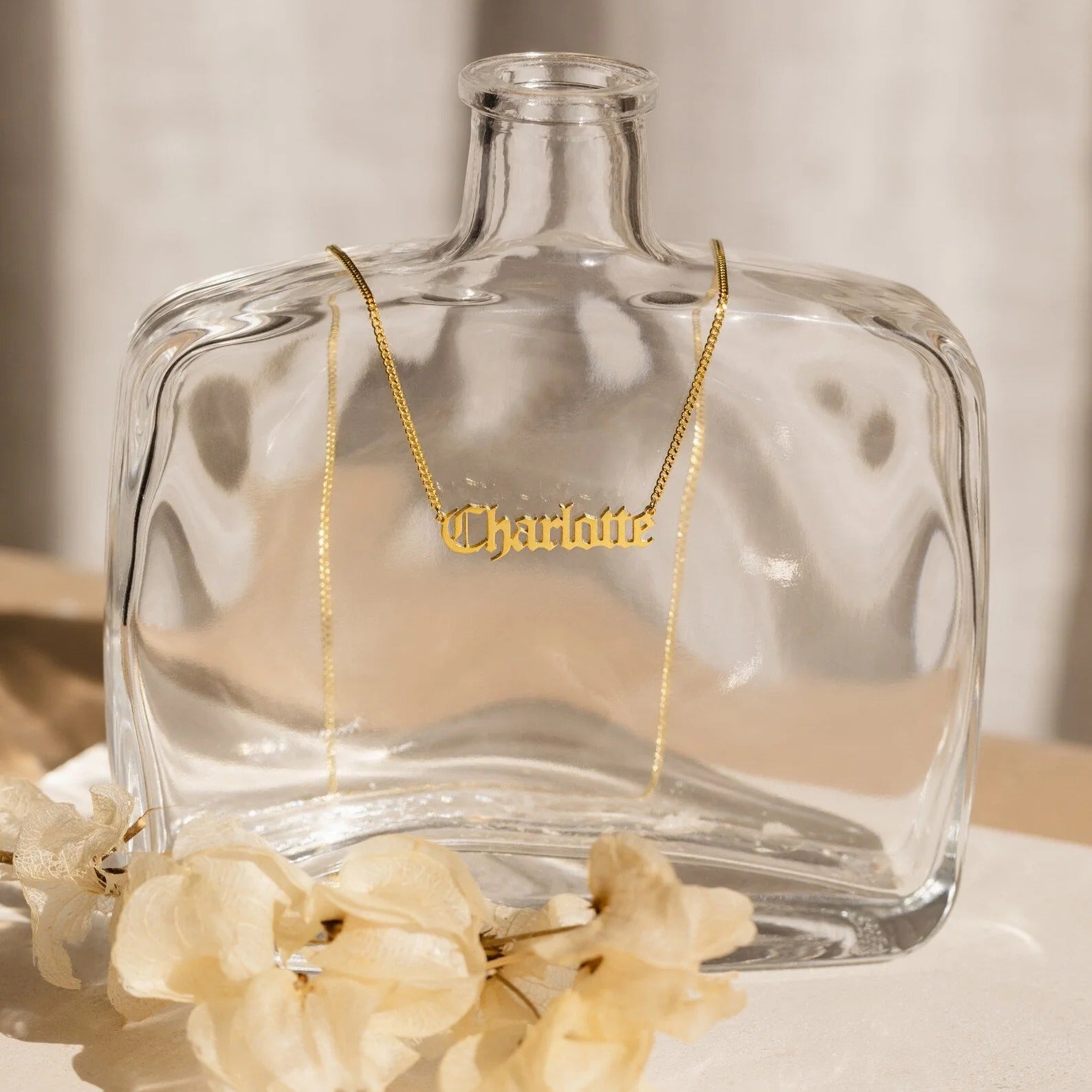 Gold necklace with 'Charlotte' pendant on a clear glass bottle with flowers at the base.
