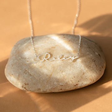 Silver necklace with 'Riley' pendant on a stone against a beige background