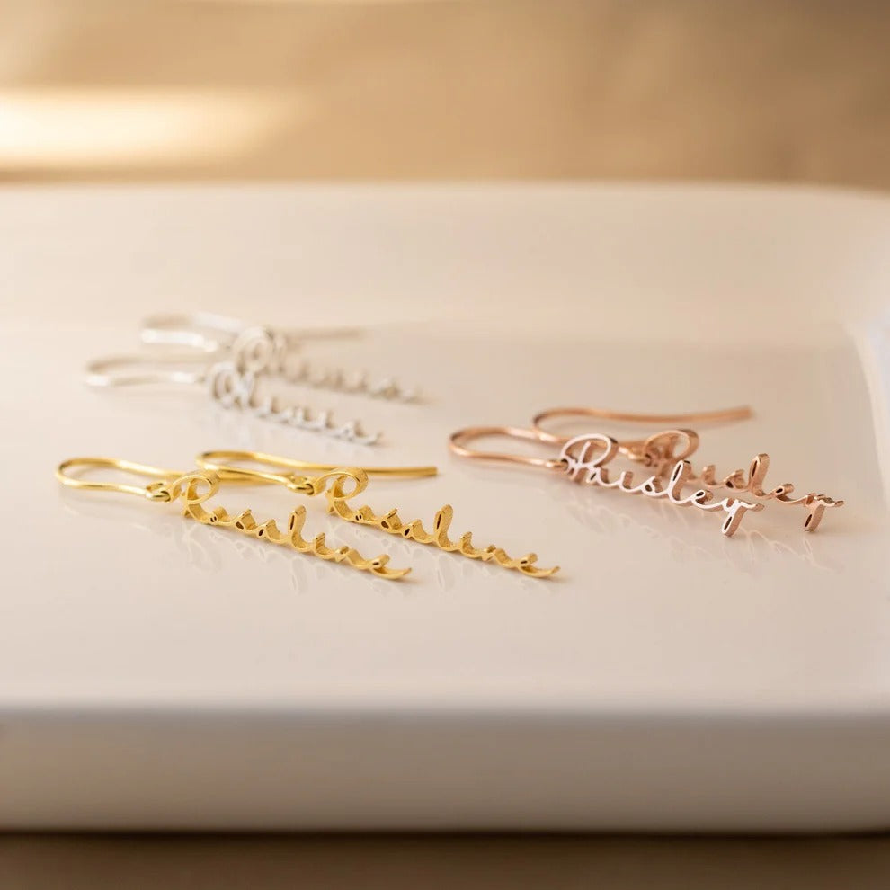 Gold and rose gold name earrings on a white surface with a beige background