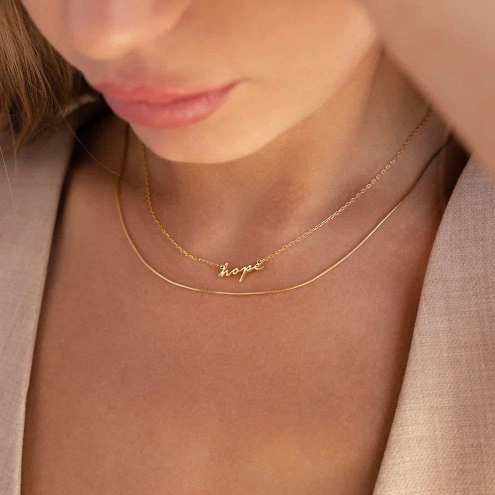 Gold necklace with 'hope' pendant on a person wearing a beige jacket.