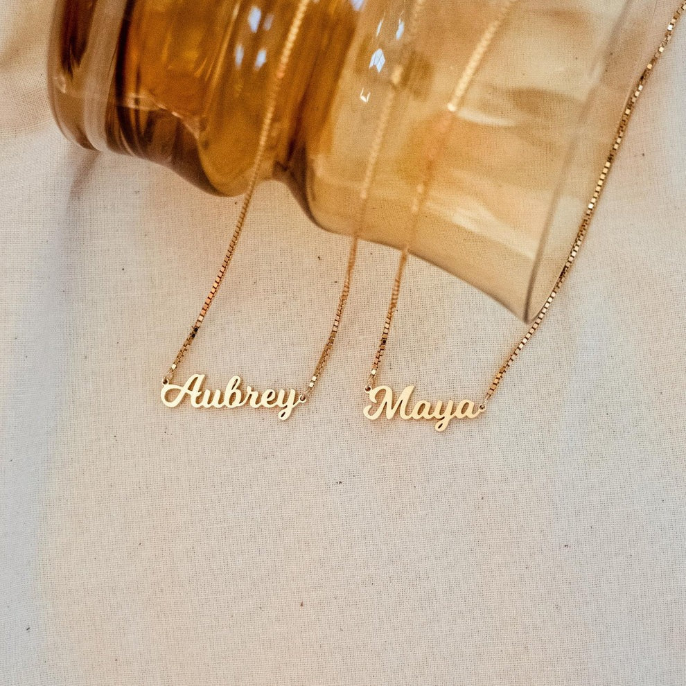 Two gold name necklaces, 'Aubrey' and 'Maya', on a beige fabric background.