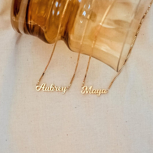 Two gold name necklaces, 'Aubrey' and 'Maya', on a beige fabric background.