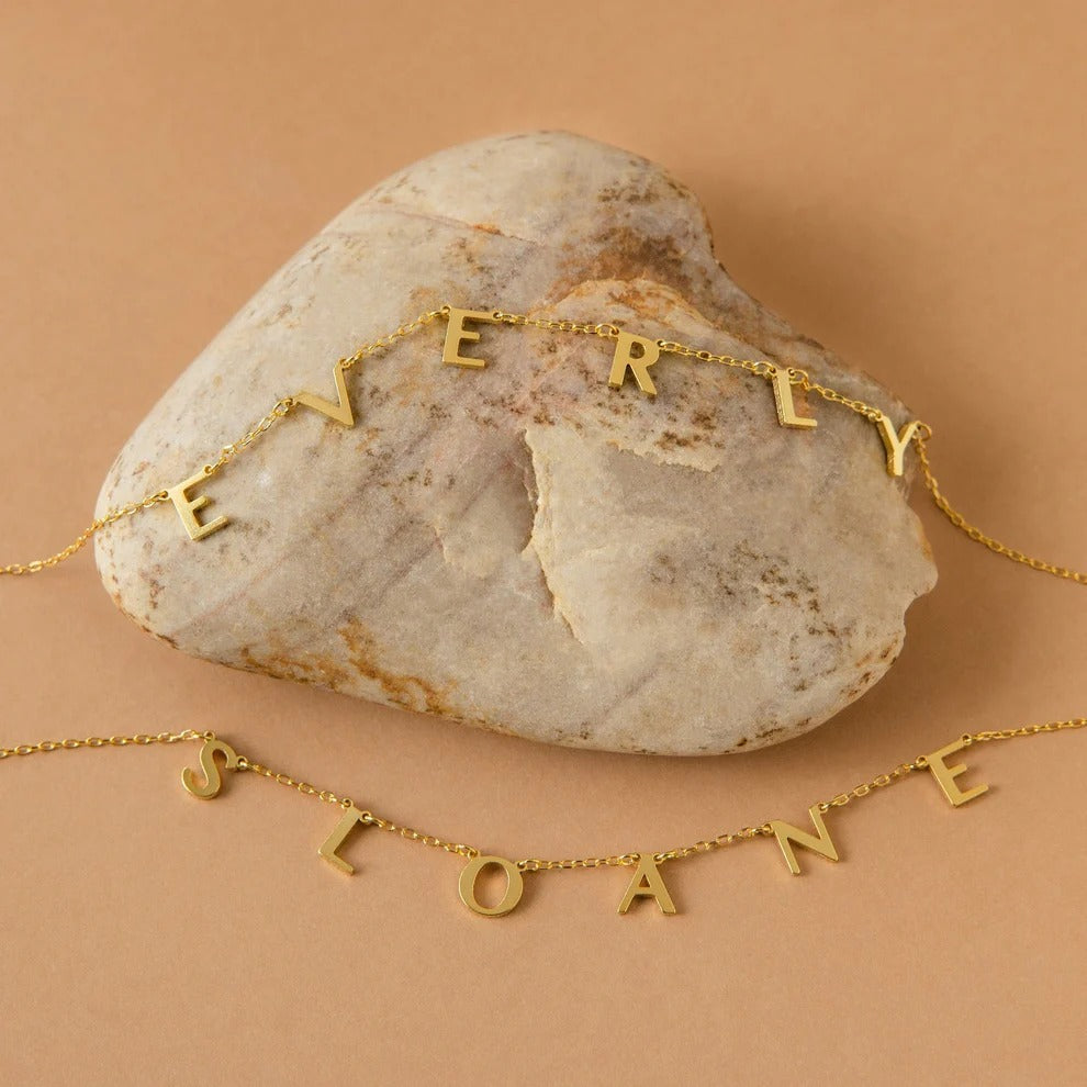 Gold necklaces with 'EVERLY' and 'SLOANE' on a stone surface