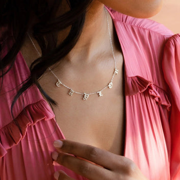 Person wearing a delicate necklace with small letters on a pink background