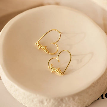 Gold heart-shaped earrings with 'Sophia' engraving on a beige stone surface