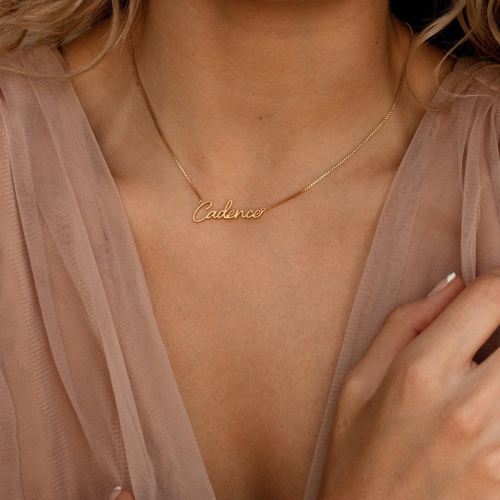 Gold necklace with 'Cadence' pendant on a person wearing a light pink top.