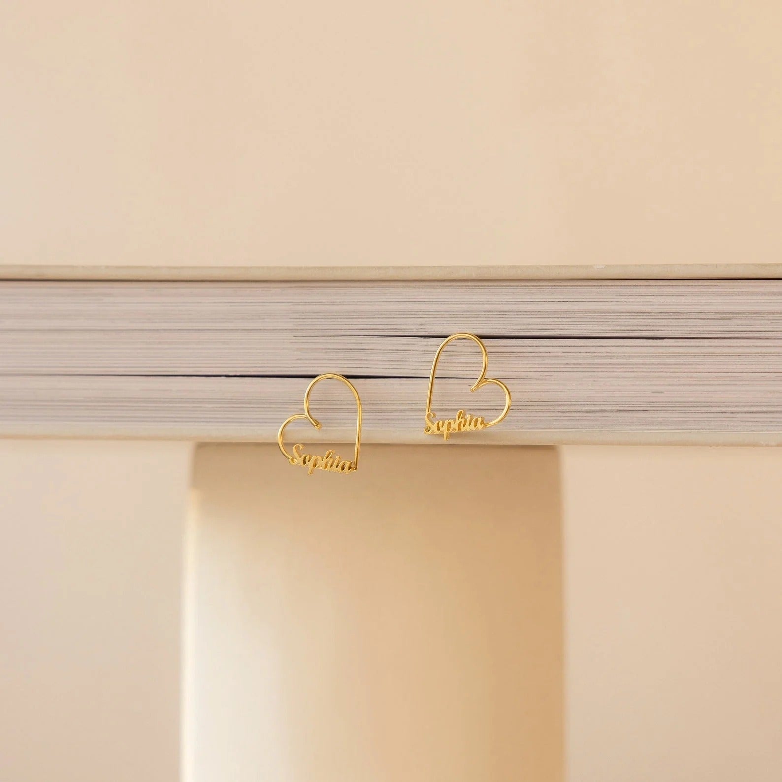 Gold heart-shaped earrings with 'Sophia' engraving on a beige background