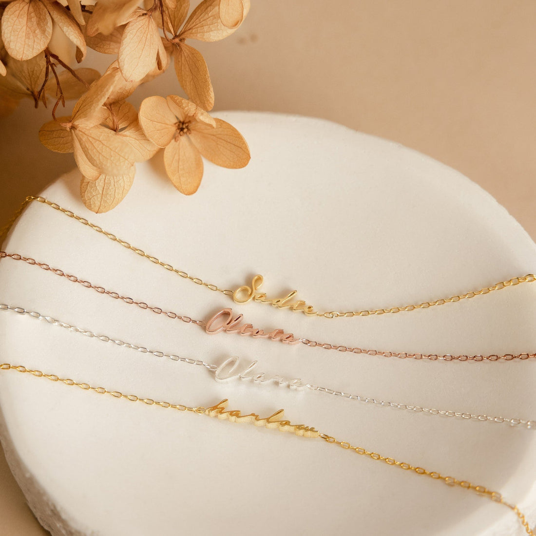 Gold necklaces with engraved names on a white surface with dried flowers.