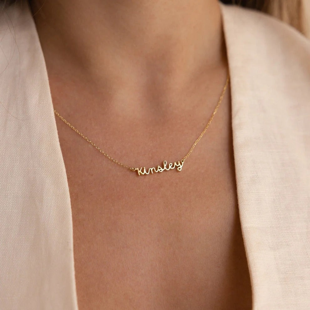 Gold necklace with 'Kinsley' pendant on a person wearing a light-colored top.