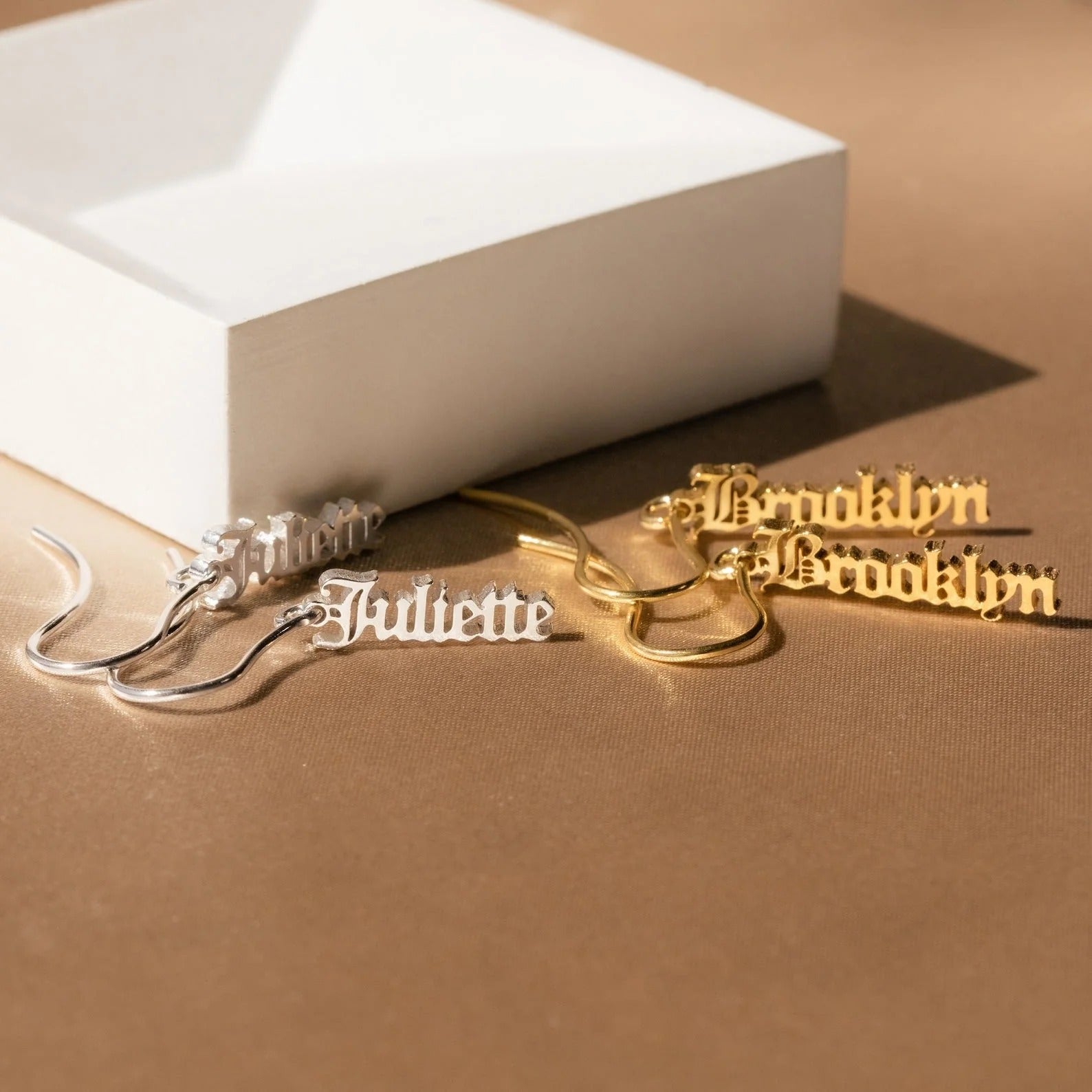 Gold and silver nameplate earrings on a brown surface with a white box.