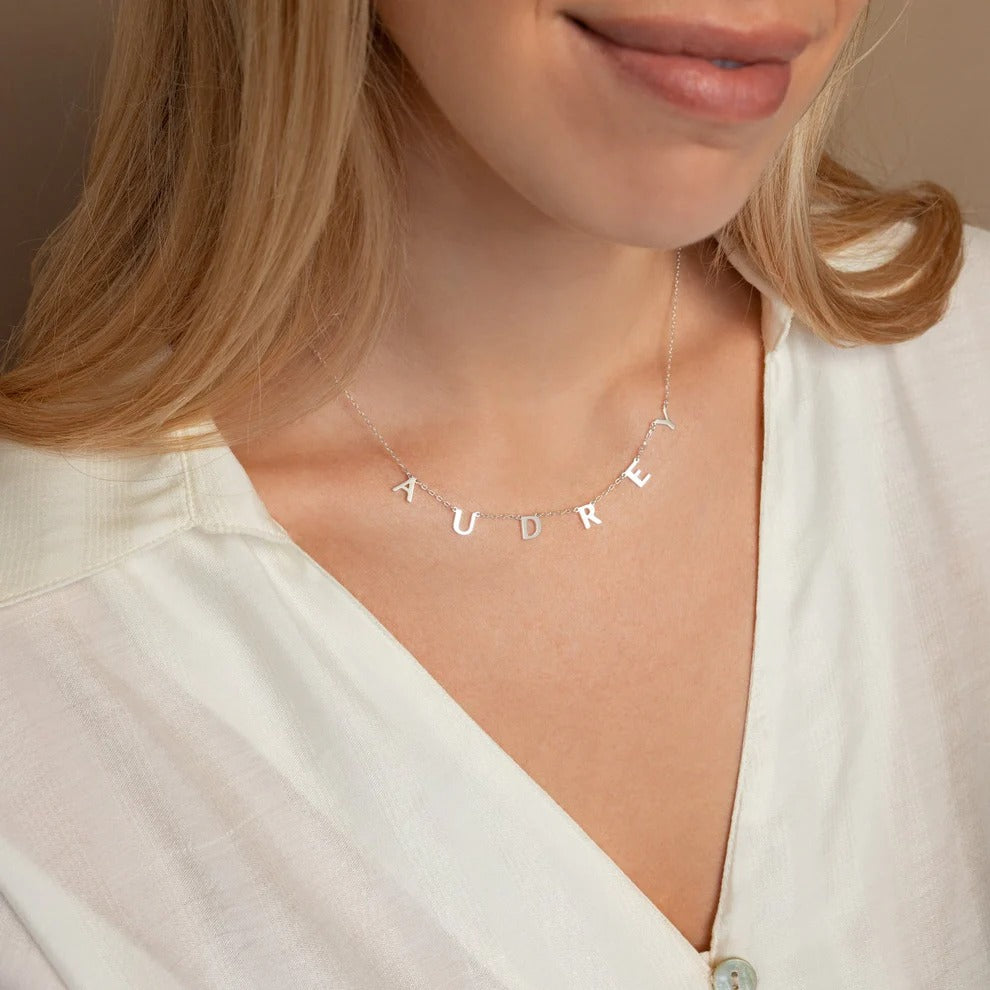Necklace with 'AUDREY' letters worn by a person in a white top.