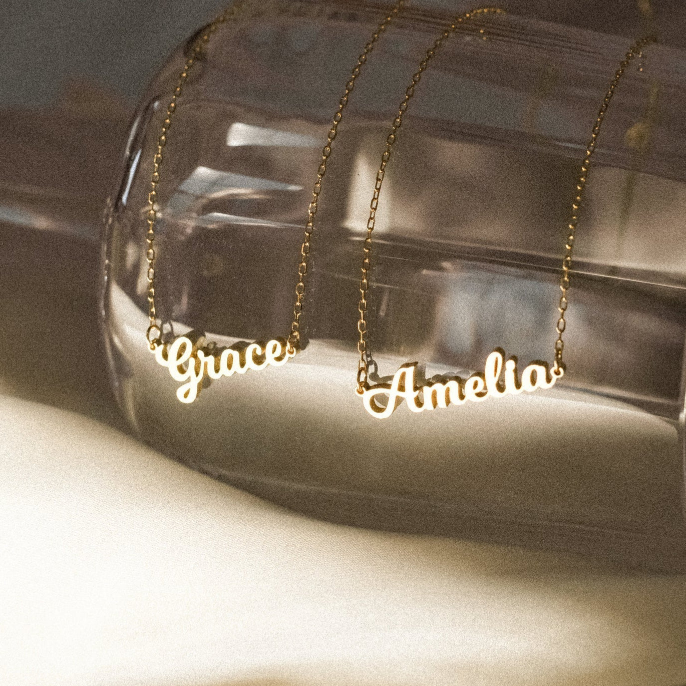 Gold name necklaces labeled 'Grace' and 'Amelia' on a clear stand.