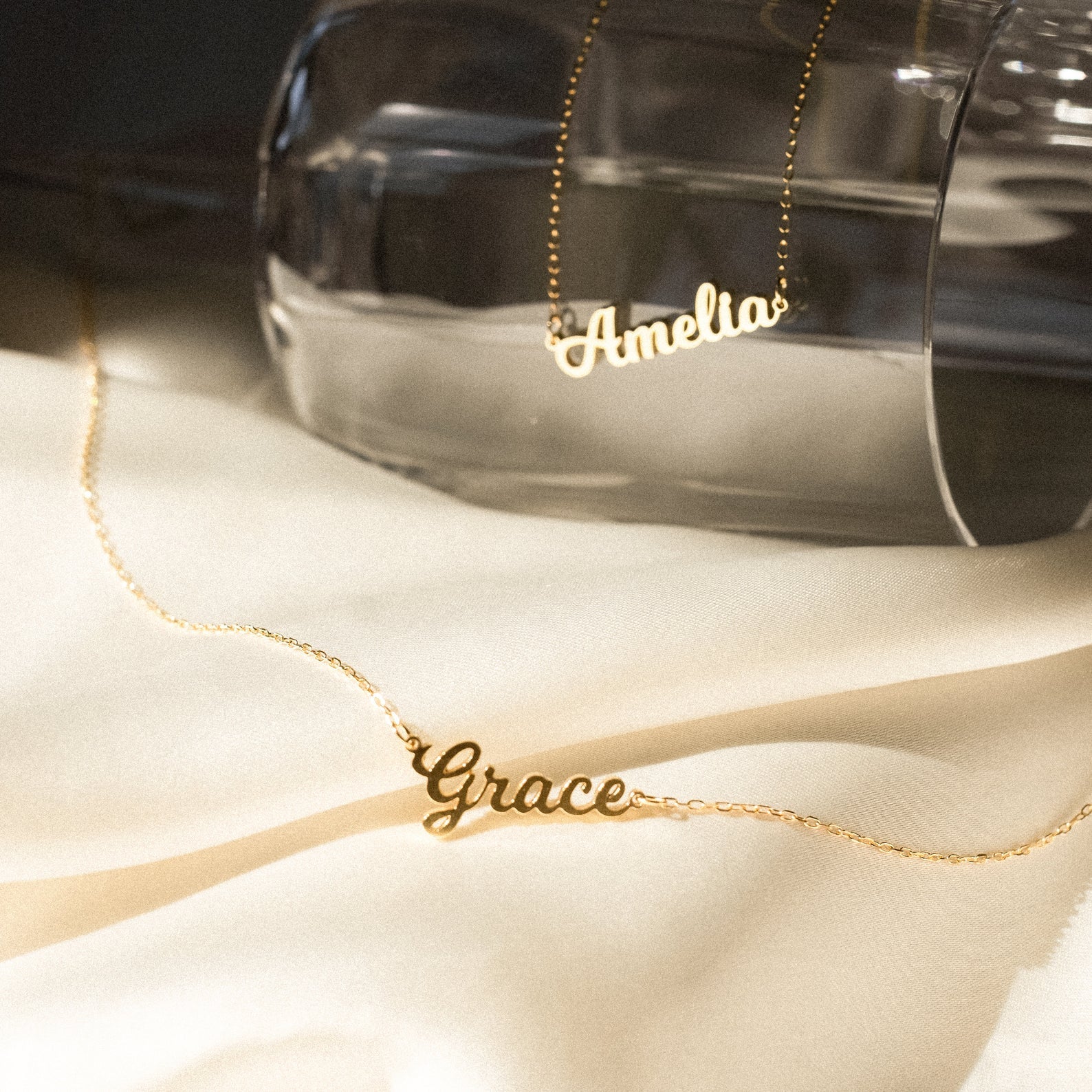 Gold name necklaces labeled 'Amelia' and 'Grace' on a white surface with a glass container in the background.