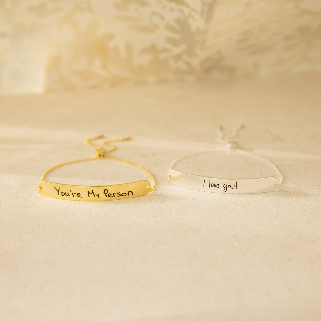 Two gold bracelets with engraved messages on a beige background