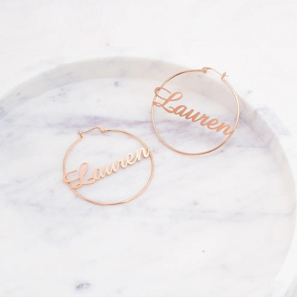 Rose gold hoop earrings with 'Lauren' engraving on a marble surface