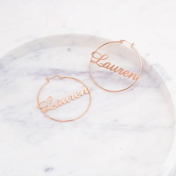 Rose gold hoop earrings with 'Lauren' engraving on a marble surface