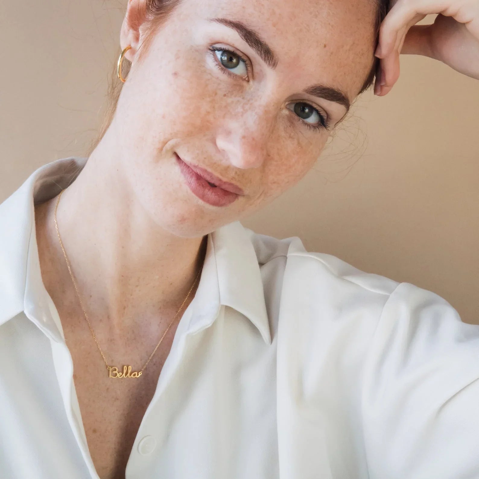 Woman wearing a gold necklace with 'Bella' pendant against a beige background