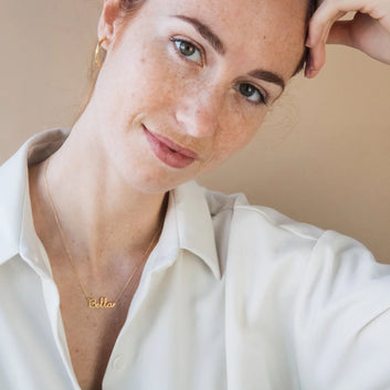 Woman wearing a gold necklace with 'Bella' pendant against a beige background