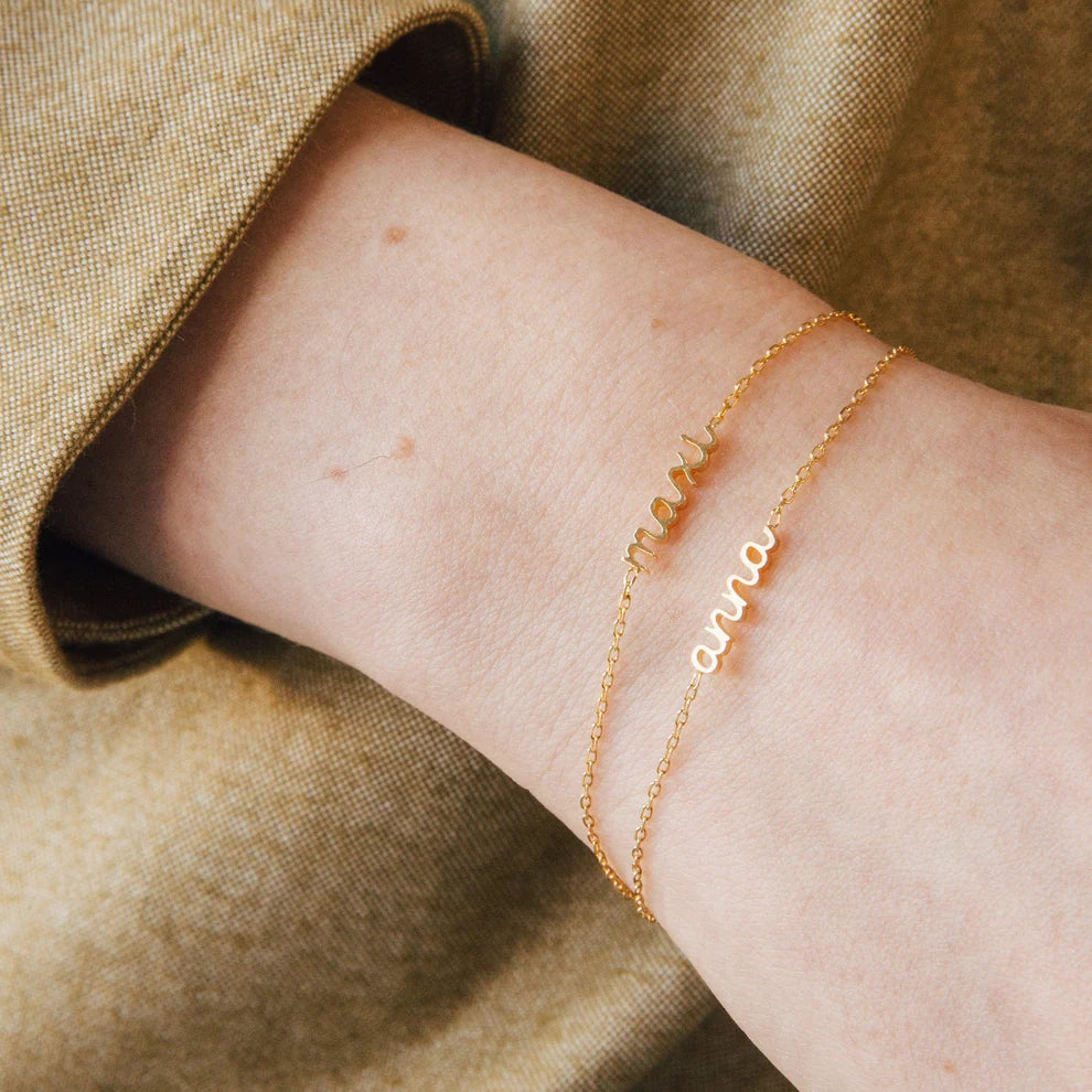Gold bracelet with engraved name on a wrist against a beige fabric background