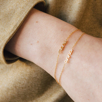 Gold bracelet with engraved name on a wrist against a beige fabric background
