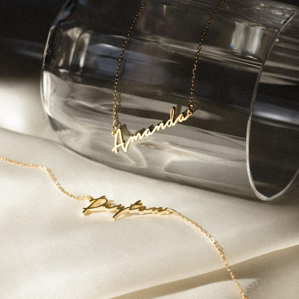 Gold name necklaces on a light surface with a clear glass object in the background