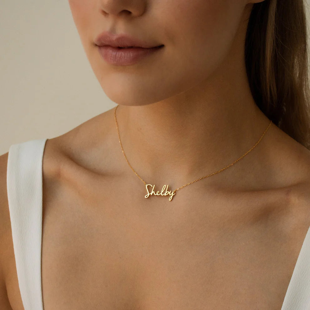 Gold necklace with 'Shelby' pendant on a person wearing a white top.