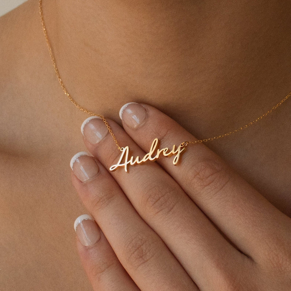Gold necklace with 'Audrey' pendant held by a hand against a skin-toned background