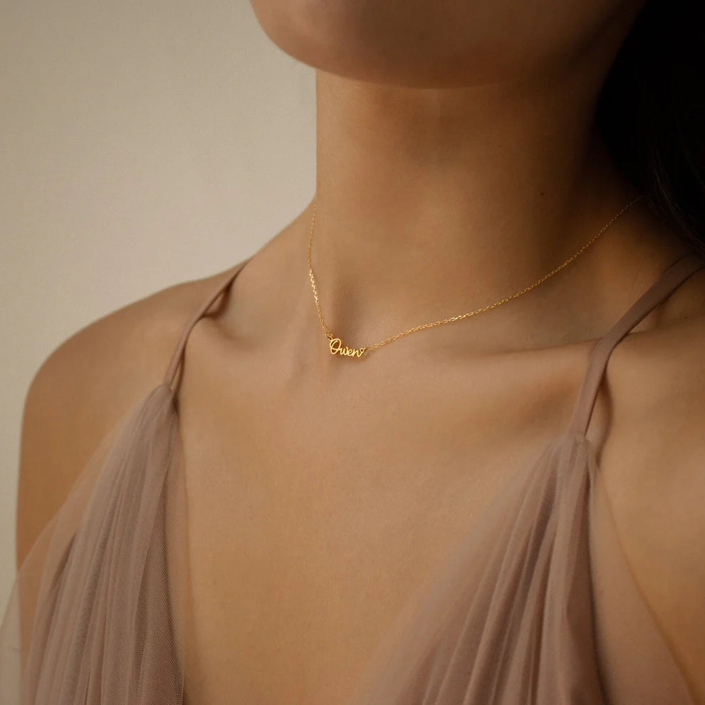 Gold necklace with a name pendant on a person wearing a beige top.
