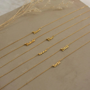 Gold name necklaces on a beige surface with dried flowers.