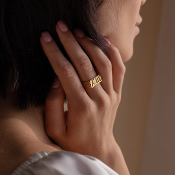 Close-up of a hand wearing a gold ring with 'Eternally' engraved, touching hair against a blurred background.