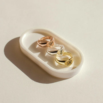Three gold rings with engraved initials on a white dish against a beige background