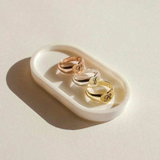 Three gold rings with engraved initials on a white dish against a beige background