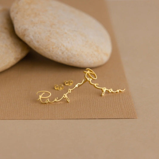 Gold script earrings on a brown surface with stones in the background