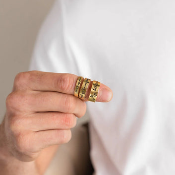 Hand wearing three gold rings with engraved text on a plain background