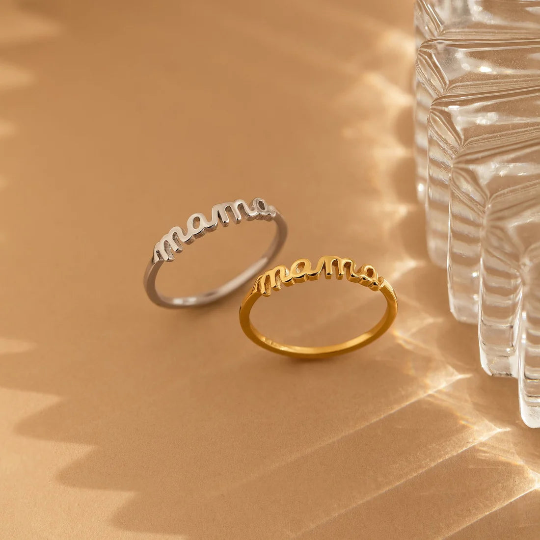 Two 'mama' rings, one gold and one silver, on a beige surface with a glass vase in the background.