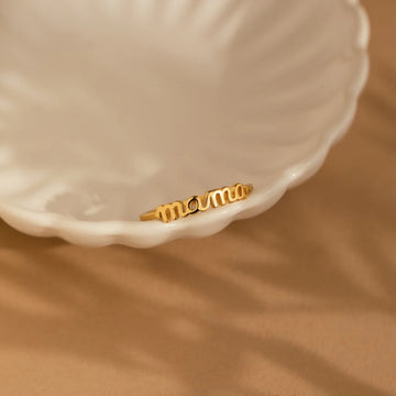 Gold 'mom' ring on a white shell against a beige background