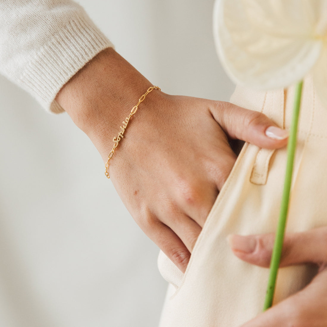 Close-up of a hand wearing a gold bracelet holding a white tulip against a neutral background