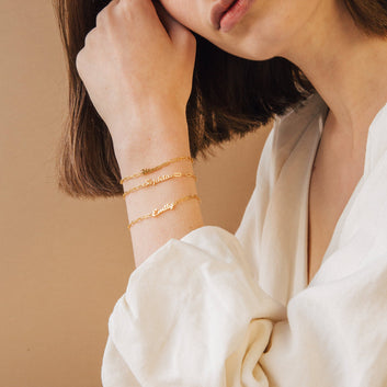 Close-up of a person wearing gold name bracelets on a beige background