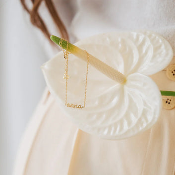 Gold necklace with 'anna' engraving on a white leaf-shaped stand.