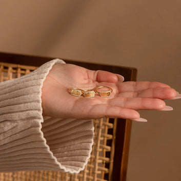 Hand wearing a beige sweater holding three gold rings against a neutral background